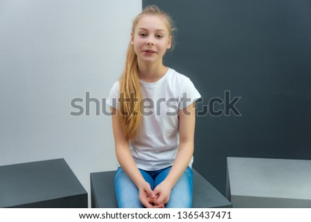 A concept portrait of a cute pretty blonde teen girl with long hair is sitting in a white T-shirt against a gray-white background and talking. She's right in front of the camera.