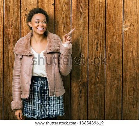 Young african american woman wearing winter coat over isolated background with a big smile on face, pointing with hand and finger to the side looking at the camera.
