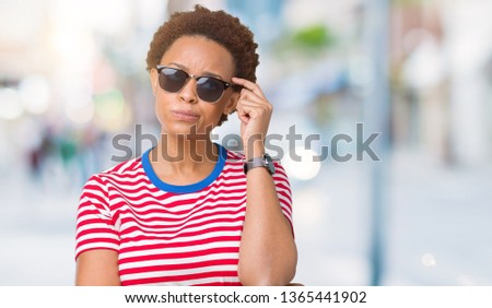 Beautiful young african american woman wearing sunglasses over isolated background with hand on chin thinking about question, pensive expression. Smiling with thoughtful face. Doubt concept.