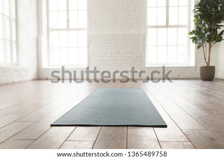 Empty room in yoga studio with unrolled yoga mat on wooden floor, modern fitness center with big windows and white brick walls, sport equipment for meditating or sport exercises close up Royalty-Free Stock Photo #1365489758