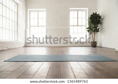 Unrolled yoga mat on wooden floor in modern fitness center or at home with big windows and white brick walls, comfortable space for doing sport exercises, meditating, yoga equipment Royalty-Free Stock Photo #1365489761