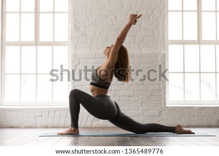 Woman in grey sportswear, bra and leggings practicing yoga, standing in anjaneyasana pose, girl doing Horse rider exercise on mat, working out at home or in yoga studio with white walls Royalty-Free Stock Photo #1365489776