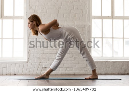 Sporty woman in white sportswear, pants and top standing in Parsvottanasana pose, doing One Sided Fold yoga exercise, attractive girl training, working out at home or in modern yoga studio side view