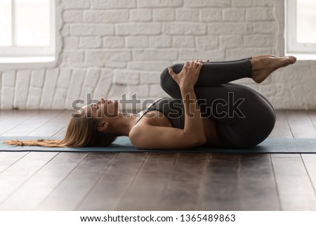 Young woman with closed eyes practicing yoga, beautiful girl in grey sportswear, pants and bra lying in Knees to Chest pose, doing Apanasana exercise, working out at home or in yoga studio
