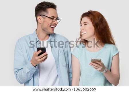 Happy millennial couple stand isolated on grey studio background hold smartphones have fun together, smiling young man and woman use cell looking at each other laughing at funny joke online