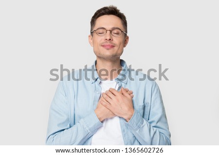 Calm young Caucasian male in glasses isolated on grey studio background hold hands on chest close to heart feel grateful, thankful European man in appreciation thanking fate. Gratitude concept