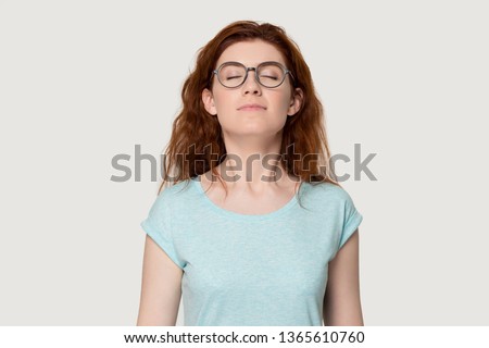 Happy young redhead woman in glasses feel calm breathe fresh air managing stress practicing exercises, peaceful red-haired girl in eyewear isolated on grey background smell inhale pleasant fragrance