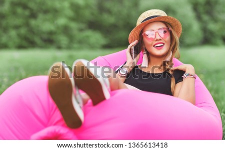 Summer lifestyle portrait of pretty smiling girl lying on the pink inflatable sofa in the park. Talking on the smartphone. Wearing stylish sunglasses, canotier. Relaxing, enjoying life on air bed