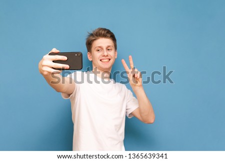 Happy guy in a white T-shirt takes a selfie on a smartphone, isolated on a blue background. Young man looks at the camera of a smartphone and poses, a selfie concept. Focus on the smartphone.