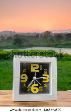 Good morning concept.  White alarm clock on wooden table over sunrise at grazing field background. Copy space