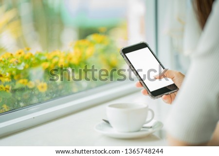 Mockup image of hand holding black mobile phone with blank white screen and hot latte coffee on white wood table in cafe