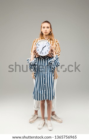 Holding round clock. Little girl in oversize striped shirt closing her face with round clock while tall friend standing behind