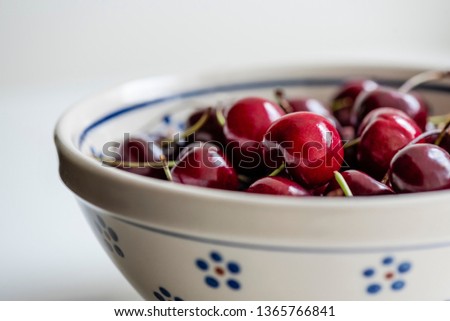 Sweet, ripe, dark red bing cherries on stems are grouped in an off-white, round Polish pottery bowl for a morning or afternoon snack. Macro, close-up photo highlighting one shiny cherry. Royalty-Free Stock Photo #1365766841