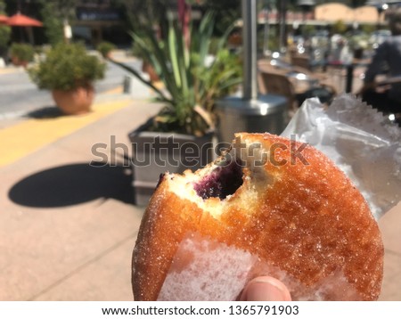 jelly donut in waxed paper with a bite missing on a sunny day with an outdoor patio behind the donut, focus on the foreground