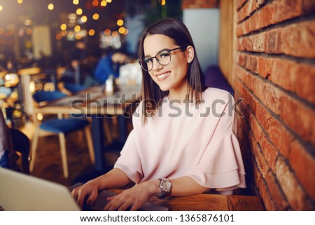 Attractive brunette with eyeglasses using laptop while sitting inn cafeteria. In background bokeh effect.