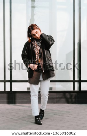 Young attractive woman wearing whie jeans and dark coat jacket posing in front of big window in the city street