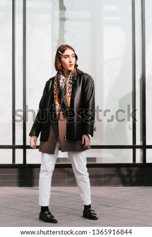 Young attractive woman wearing whie jeans and dark coat jacket posing in front of big window in the city street