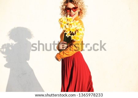 young curly blond  woman holding yellow flowers, wearing turtleneck, red lipstick, skirt and sunglasses against the white wall