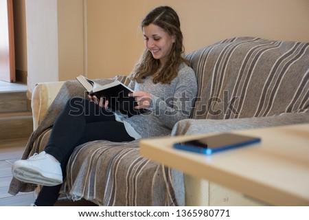 Young caucasian girl reads a book on the sofa leaving smartphone on the table. I love book concept