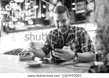 Smiling handsome Caucasian man in plaid shirt using smart phone and drinking coffee. Selective focus on hands, cafe interior. Black and white photo.