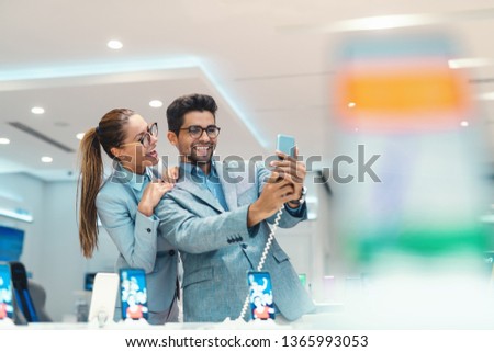 Young attractive multicultural couple dressed elegant taking selfie with new smart phone in tech store.