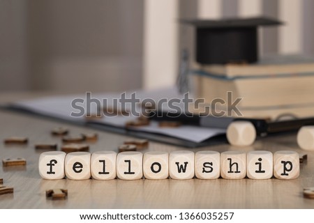 Word FELLOWSHIP composed of wooden dices. Black graduate hat and books in the background. Closeup Royalty-Free Stock Photo #1366035257