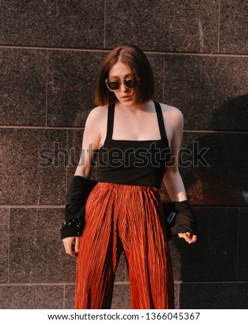 Model girl wearing dark jacket and black top and red pants and sunglasses posing in front of dark wall of modern building.