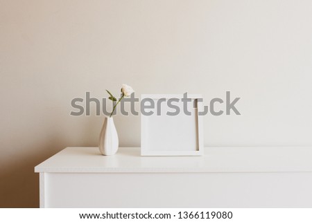 Close up of single rose in small white vase next to blank square picture frame on sideboard against neutral wall - warm matte filter effect