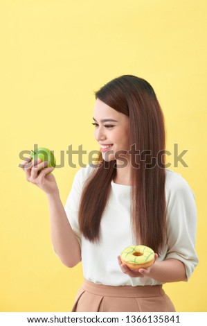 Portrait of a smiling young asian woman choosing between donut and green apple isolated over yellow background