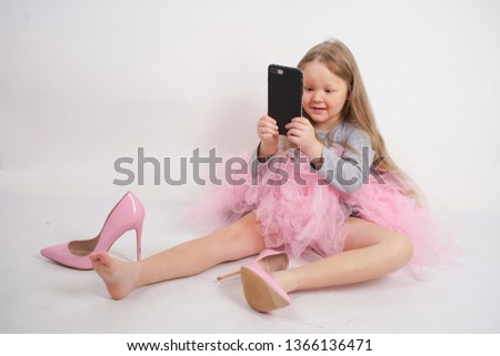 a little caucasian child girl sits and puts mother's high-heeled pink shoes, taking pictures of it all and selfies on her smartphone on white studio background