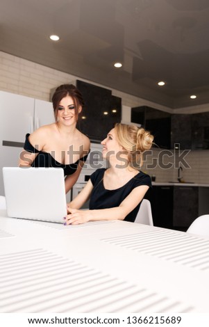 Two young white women in the kitchen with a laptop, in black dresses with makeup and hair. Model plus size