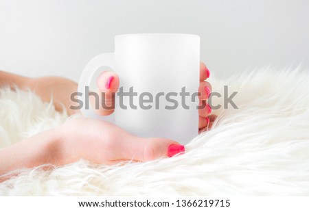 Blank frosted coffee mug held in hands on sheepskin rug, drinkware mock up