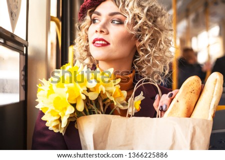 portrait young fashion woman with curl blond hair, beautiful make up and red lipstick, siting at the bus , wearing yellow blouse , beret , grey bag and coat. holding a flowers
