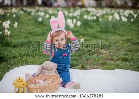 Easter. Adorable little blue eyed girl with pink rabbit ears is sitting in green meadow near blooming daffodils. Spring Holidays.