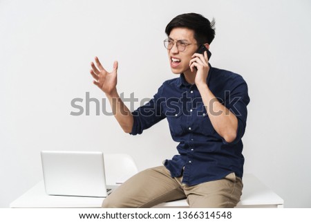 Photo of nervous asian businessman 20s wearing eyeglasses talking on smartphone while working with laptop in office isolated over white background