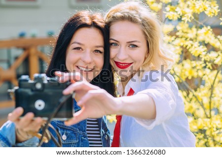 Outdoor portraits of two pretty girls friend taking selfie on vintage camera, enjoy nice day, looking on camera and making funny faces over yelow blossom tree. Travel, lifestyle concept.