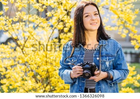 Portrait of a positive smiling caucasian hipster woman in blue jeans jacket taking picture with an old-fashion camera looking at camera outdoor with yellow blossom tree on background.