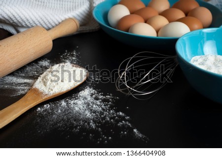 Preparation of the dough. Flour, eggs, whisk, rolling pin and a kitchen towel on the black table. A bunch of flour on a wooden spoon and lots of eggs on the blue plate. Baking reciepe. Close up. 