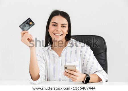 Photo of successful brunette businesswoman 30s holding smartphone and plastic credit card while sitting in armchair at table in office isolated over white background