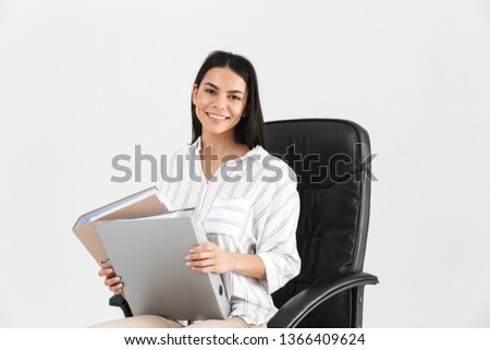 Photo of caucasian brunette businesswoman 30s smiling and holding bunch of paper folders while sitting in black armchair in office isolated over white background
