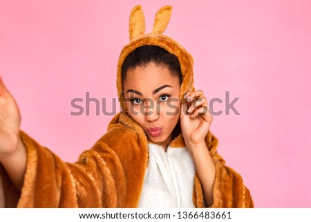 Young woman in bunny kigurumi standing isolated on pink background taking selfie picture on smartphone pouting lips to camera cute close-up