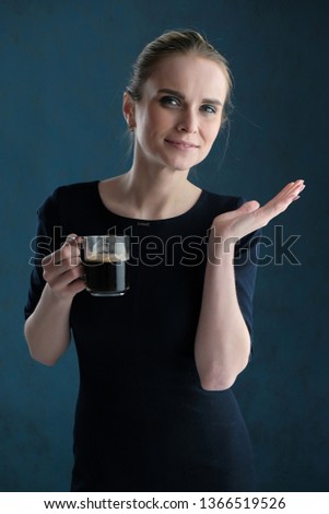 Studio portrait concept of a beautiful fashionable blonde girl standing in a business dress on a blue background drinking coffee. She is right in front of the camera, smiling.