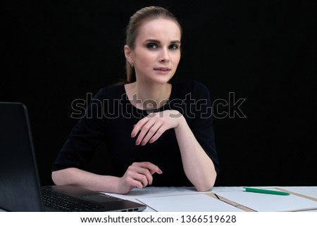 Studio portrait concept of a beautiful fashionable blonde girl and talking in a business dress on black background sitting with a laptop. She is right in front of the camera, smiling.