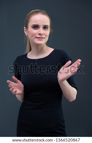 Studio portrait concept of a beautiful fashionable blond business girl standing and talking in a business dress against a gray background. She is standing right in front of the camera and smiling.