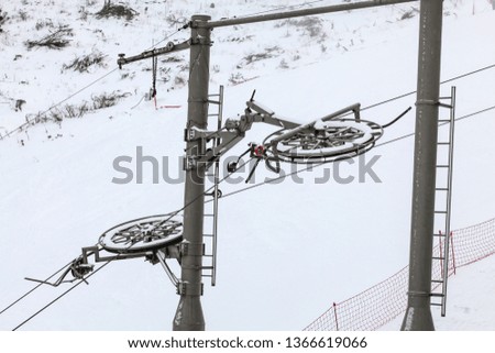Large metal wheels for steel cables on top of ski lift supporting pillar, snow covered piste in background