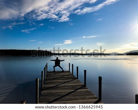 A girl that is doing a yoga pose on a dock on a lake in front of the mountains.