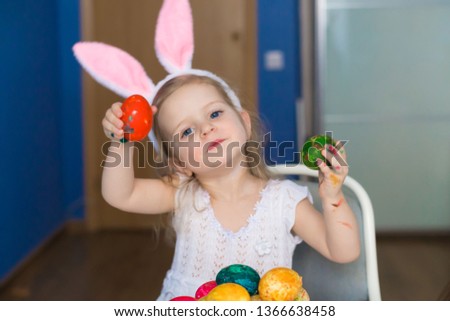 Beautiful little blonde girl, has happy fun smiling face, pretty eyes, white t-shirt, hare ears, paint easter eggs. Child portrait and kids hobby concept. Holiday accessories. 