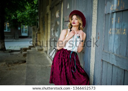 Fashionable and beautiful blonde model girl in stylish red velvet velour skirt, white blouse and hat, posed with phone and earphones.