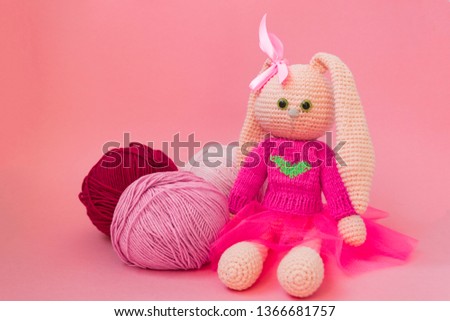 Knitted pink rabbit handmade. Easter bunny, in a pink knitted sweater and in a pink skirt. Sits among balls of yarn and pink flowers, On karalovom background.