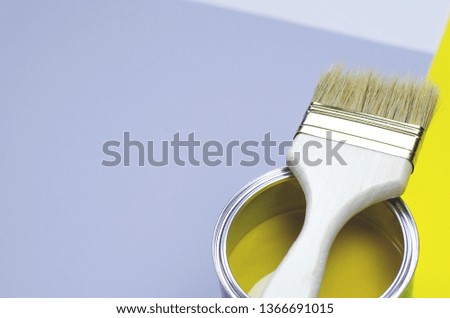 A can of yellow paint with a brush on a gray-yellow with a white background.